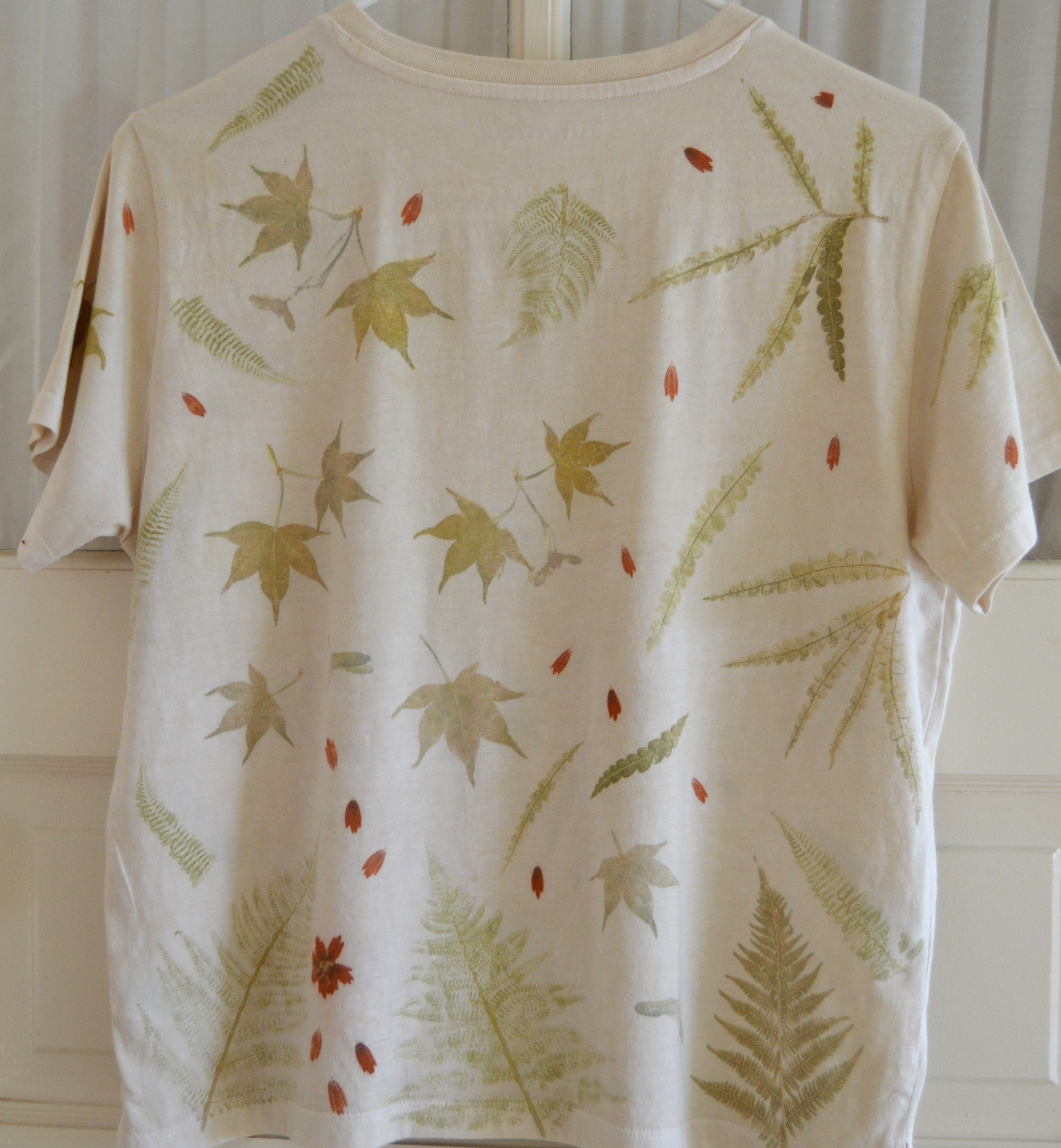 Back of a cream combed-cotton T-shirt pounded with late-summer ferns, maple leaves, and red blooms — mirror image of the front